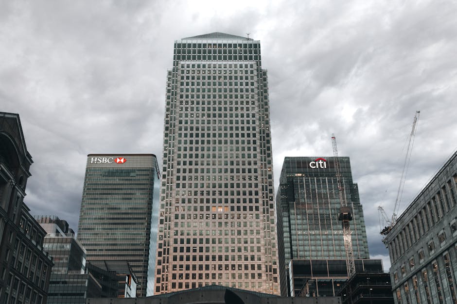 A cityscape featuring a central high-rise office building with a grid-like facade and multiple illuminated windows, surrounded by other tall buildings, including HSBC and Citi bank headquarters, with 