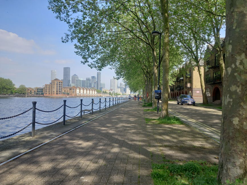 A wide shot of the Canary Wharf business district during dusk, featuring tall modern office buildings with glass facades illuminated from within. The buildings are positioned along a waterway, with so